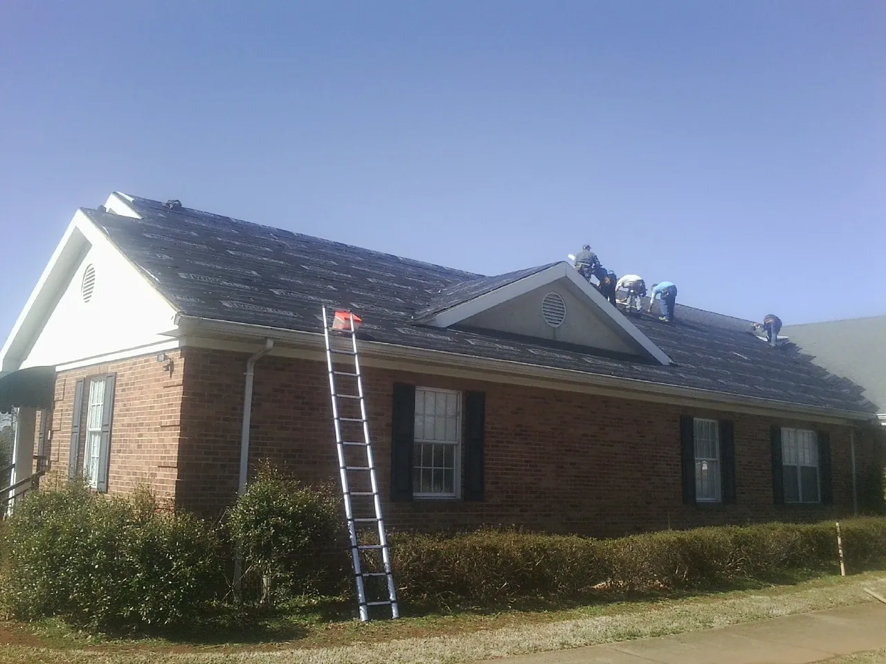 Crew tearing shingles off a brick ranch roof with a ladder set — Metal Roof Repair work by our crew in Timnath