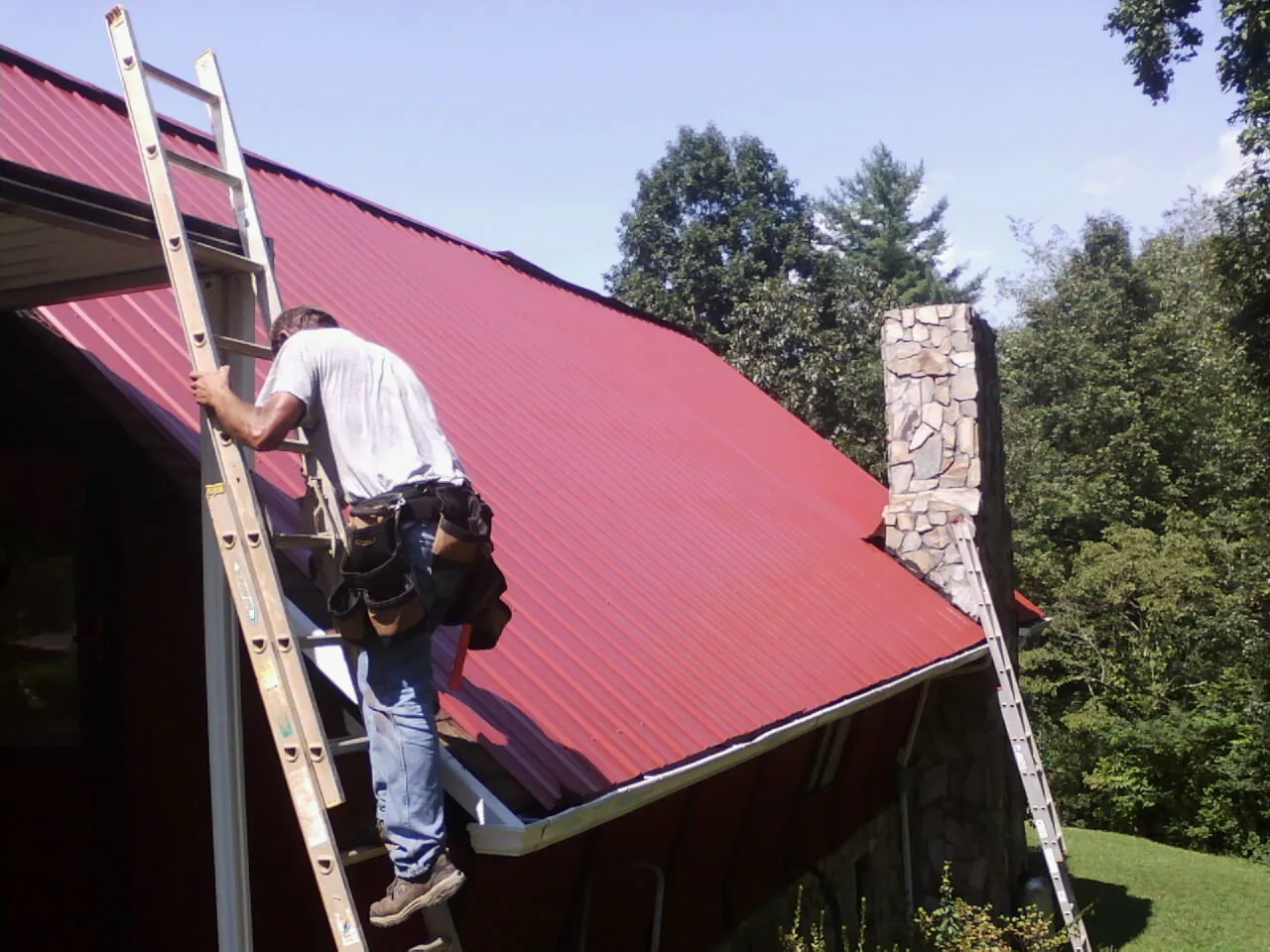 Roofer on a ladder beside a red metal roof and stone chimney — Soffit & Fascia Repair by Roofing Brothers in Timnath